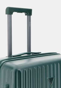 Valigia rigida verde con scanalature verticali, dotata di manico telescopico grigio e un logo nell'angolo in basso.