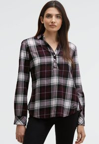 Woman with long brown hair wearing a purple and white plaid buttoned shirt and black pants against a plain light background.