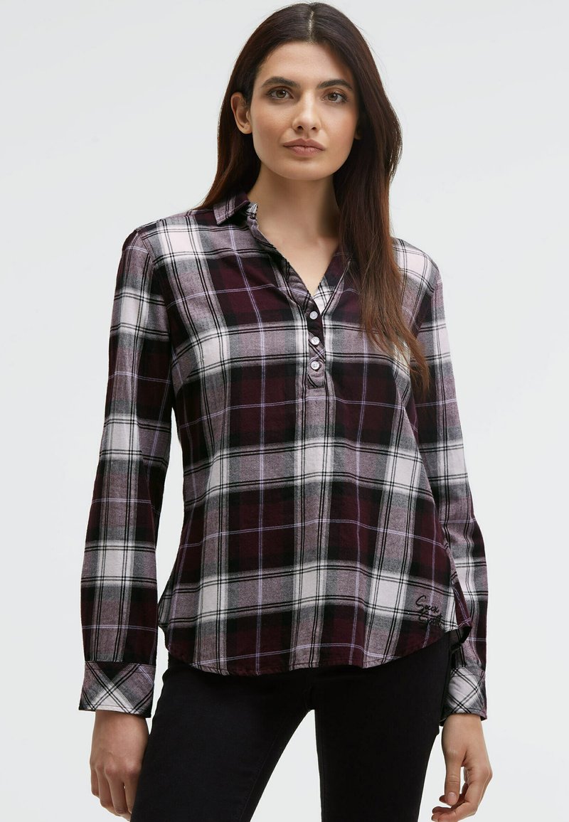 Woman with long brown hair wearing a purple and white plaid buttoned shirt and black pants against a plain light background.