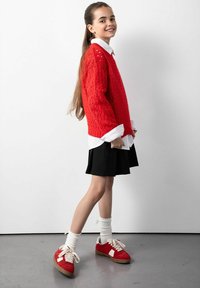 Czerwony sweter z dzianiny o luźnym kroju, biały sweter z kołnierzykiem pod spodem, czarna spódnica oraz czerwone sneakersy z białymi akcentami i kremowe skarpetki.