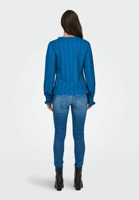 Blouse bleue texturée avec un col à volants et des manches longues, associée à un jeans bleu ajusté et des bottes noires à talons, vue de dos.