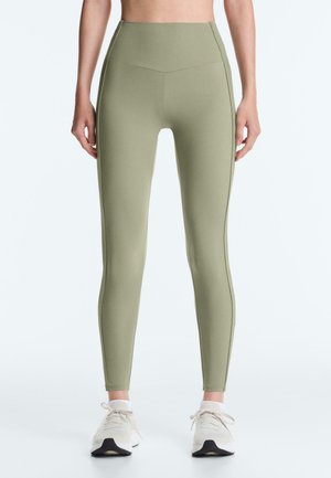 Osoba nosiaca olivovo zelené legíny s vysokým pásom a biele športové topánky, stojaca vzpriamene pred jednoliatym pozadím.