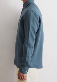 Kragad långärmad skjorta i ett mjukt, texturerat blått tyg, med en slim fit och beige knappar längs placket och ärmsluten.