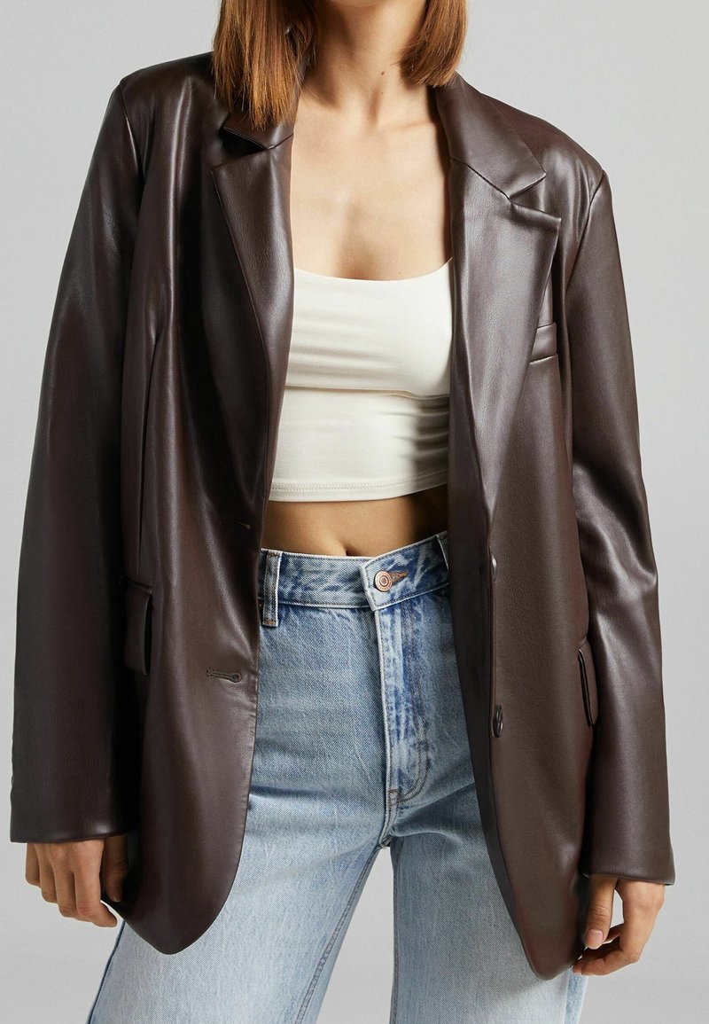 Femme portant un blazer en cuir marron foncé surdimensionné, un débardeur blanc court, et un jean taille haute bleu clair sur un fond uni.