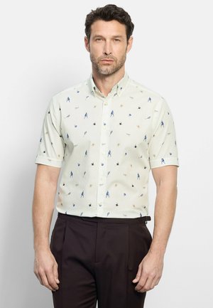 Homme portant une chemise légère à manches courtes avec des motifs sportifs bleus et un pantalon foncé, debout devant un fond uni.