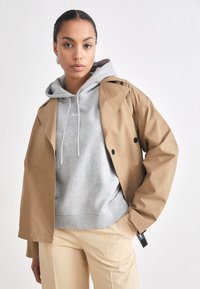Un trench coat maro peste un hanorac gri deschis cu șnururi contrastante, având o croială lejeră și pantaloni bej.
