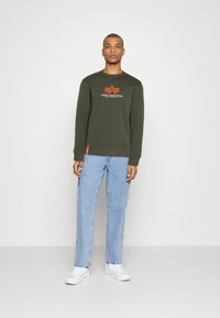 Sudadera verde con cuello redondo y logotipo de Alpha Industries; combinada con jeans rectos de color azul claro y zapatillas blancas.