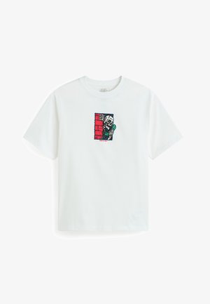 Maglietta bianca a maniche corte con grafica di uno scheletro che tiene un telefono verde, che sbircia da dietro un muro di mattoni rossi, con la scritta "Mi sento così vivo."