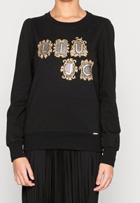 Sudadera negra con diseños gráficos ornamentados en dorado y cuadros. Cuello redondo clásico con mangas abullonadas, confeccionada en tela suave.