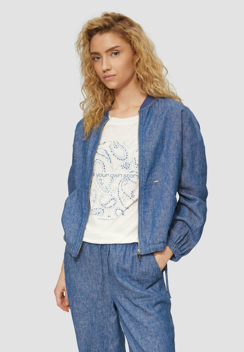 Blauwe denim bomberjack met een rits aan de voorkant, geribbelde kraag enGathered mouwen, gedragen over een licht T-shirt met een patronenontwerp.