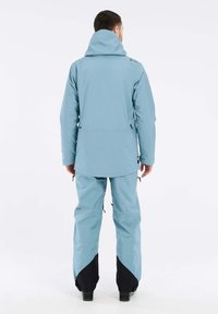 Lichtblauwe skijack met capuchon, met een langere achterkant en ritszakken, in combinatie met bijpassende lichtblauwe broeken met zwarte accenten.