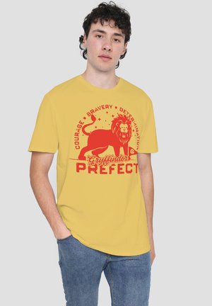 Young man wearing a yellow T-shirt with a red Gryffindor lion and text "Courage, Bravery, Determination, Gryffindor Prefect," paired with blue jeans.
