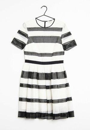 Robe blanche à manches courtes avec des bandes horizontales en dentelle noire, taille cintrée et encolure ronde, suspendue à un cintre noir sur un fond blanc.