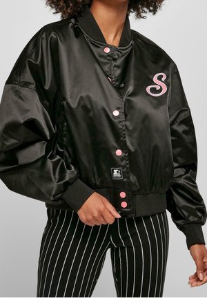Mujer con chaqueta bomber negra de satén con botones rosas y emblema de "S", combinada con pantalones a rayas verticales en blanco y negro.