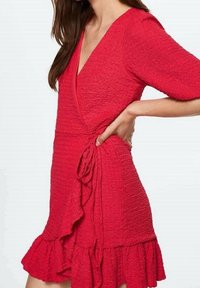 Czerwona sukienka kopertowa z fakturowanej tkaniny, dekoltem w serek, rękawami o długości trzech ćwiartek i falbaną u dołu. Posiada detal w postaci związanej talii.