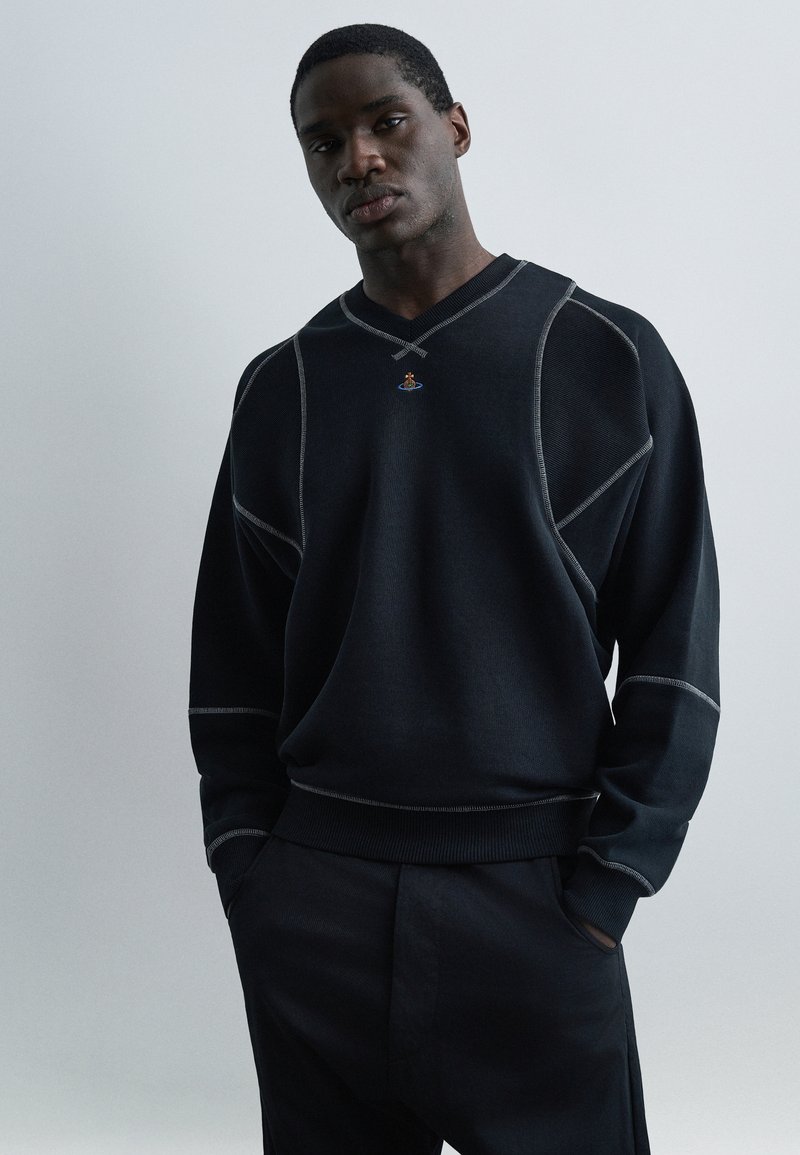 Νεαρός άνδρας με τα χέρια στις τσέπες, φορώντας μαύρο φούτερ με αντίθετες ραφές και μικρό πολύχρωμο έμβλημα στο στήθος, σε απλό φόντο.