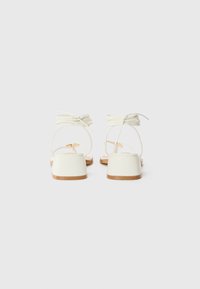 White block-heeled sandals with thin ankle straps, featuring a smooth finish and a rounded toe design. Back view shows a minimalist style.