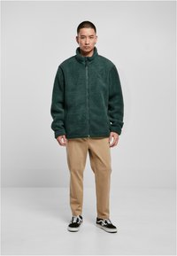 Giacca in pile con zip di colore verde scuro, caratterizzata da un colletto alto, una vestibilità rilassata e una texture morbida. Indossata sopra pantaloni color khaki e sneakers nere.