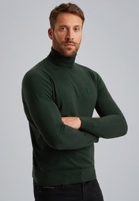 Dunkelgrüner Rollkragenpullover aus glattem Strickstoff, mit taillierter Passform und einem kleinen, rechteckigen Logo-Patch auf der Brust.
