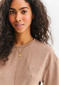 Kobieta o kręconych ciemnych włosach, ubrana w jasnobrązową koszulę z haftowanym wzorem rzęsy oraz złoty naszyjnik z wisiorkiem w kształcie kompasu.