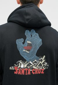 Crna dukserica s grafičkim dizajnom plave, žičane ruke s otvorenim ustima, postavljena na izlizanu pozadinu s natpisom "SANTA CRUZ."