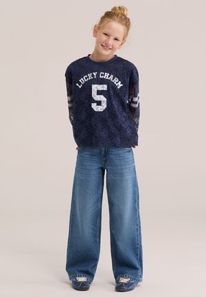 Jeune fille souriante aux cheveux blonds en chignon porte un haut en dentelle bleu marine "Lucky Charm", un jean bleu à jambes larges et des baskets bleu marine, debout devant un fond uni.