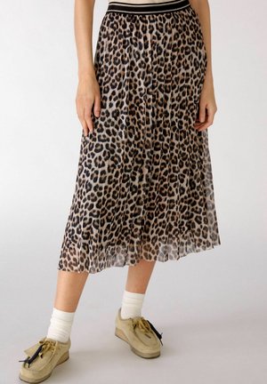 Kobieta w plisowanej spódnicy midi w panterkę, białych skarpetkach i beżowych mokasynach stoi na jednolitym tle.