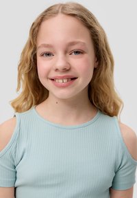 Fille blonde souriante avec des cheveux ondulés portant un haut côtelé bleu clair à épaules dénudées, sur un fond léger et uni gris.