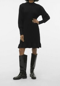 Robe en tricot noire à manches longues et texture côtelée, avec un ourlet évasé. Assortie à des bottes noires brillantes montant jusqu'au genou.