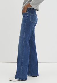 Jean en denim à jambes larges et coupe droite, de couleur bleu foncé, avec poches avant, poches arrière et détails de couture subtils. Texture lisse.