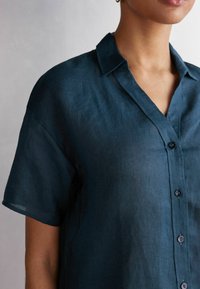 Camisa de lino verde oscuro con mangas cortas, cuello con muesca y botones en la parte frontal. La tela tiene un aspecto ligeramente texturizado.