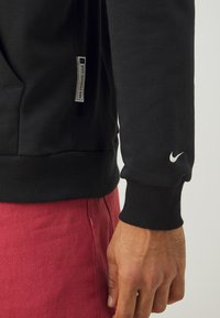 Sudadera negra con puños acanalados, que presenta un pequeño logo blanco de Nike en la manga. La etiqueta lateral dice "Nike Standard Issue". Pantalones rojos visibles.