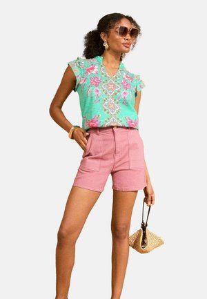 Floral-patterned green blouse with short frilled sleeves, paired with pink high-waisted shorts. Accessories include large sunglasses and a woven bag.