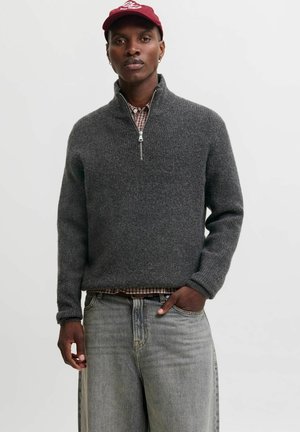 Homme portant un pull gris foncé à demi-fermeture éclair sur une chemise à carreaux, un jean gris clair et une casquette de baseball rouge, debout devant un fond uni.