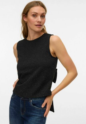 Sieviete tērpusies bezroku melnā teksturētā topā ar sānu loku un zilās džinsās, pozējot ar vienu roku kabatā pret balto fona.