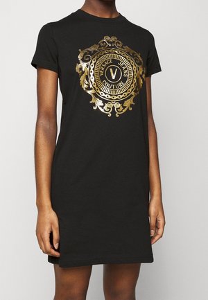 Zwarte jurk met korte mouwen, met een gouden sierlijk cirkelvormig embleem en de tekst "Versace Jeans Couture", gedragen door een staande persoon, tot halverwege de dij.