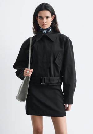 Femme portant une veste noire ceinturée et une jupe, portant un sac à bandoulière gris, avec un foulard fleuri autour du cou et des boucles d'oreilles créoles argentées.