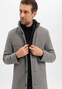 Grijze visgraatmantel met een hoge kraag, knoopsluiting aan de voorkant en twee zakken, gedragen over een zwarte coltrui, met een gladde textuur.