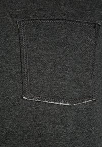 Dunkelgrauer Baumwollstoff mit einer gesteppten Tasche. Die Kanten der Tasche zeigen einen ausgefransten Detail. Glatte Textur mit einem subtilen Melange-Muster.