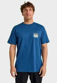 Homem com cabelo encaracolado curto, usando uma t-shirt azul com uma pequena ilustração colorida de um rio e sol no lado esquerdo do peito.