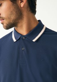 Tumesinist polo-särki tekstureeritud kangast, kaelusega, millel on kaks valget triipu ja väike tikitud logo rinnal.