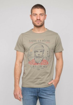 T-shirt à manches courtes kaki avec un imprimé graphique représentant un homme barbu portant un bonnet. Doté d'un texte rouge et noir, d'un col rond et d'une coupe décontractée.