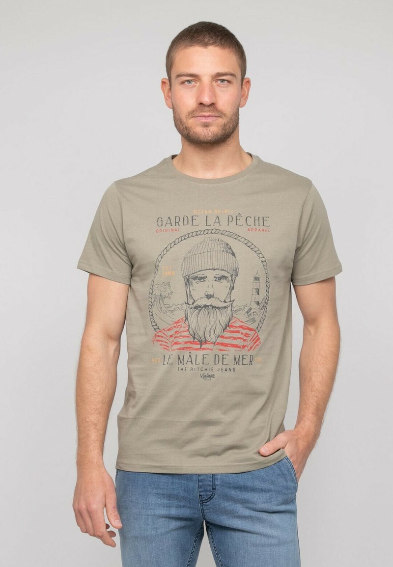 T-shirt à manches courtes kaki avec un imprimé graphique représentant un homme barbu portant un bonnet. Doté d'un texte rouge et noir, d'un col rond et d'une coupe décontractée.