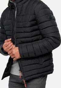 Schwarze Pufferjacke mit horizontaler Steppung, hohem Kragen und Reißverschluss. Mit orangen Zipper-Anhängern und Logo-Patch am Ärmel.