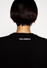 Černé bavlněné tričko s kulatým výstřihem. Na zadní straně se nachází bílý nápis "KARL LAGERFELD" poblíž výstřihu. Hladký povrch.