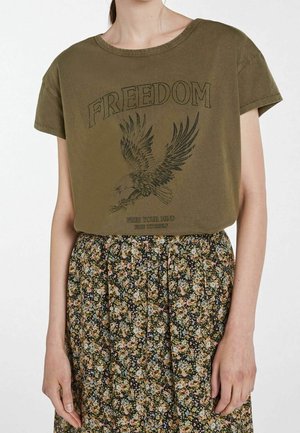 Camiseta de algodón verde oliva con cuello redondo y mangas cortas, con un gráfico de un águila negra y el texto "LIBERTAD". Combinada con una falda floral.