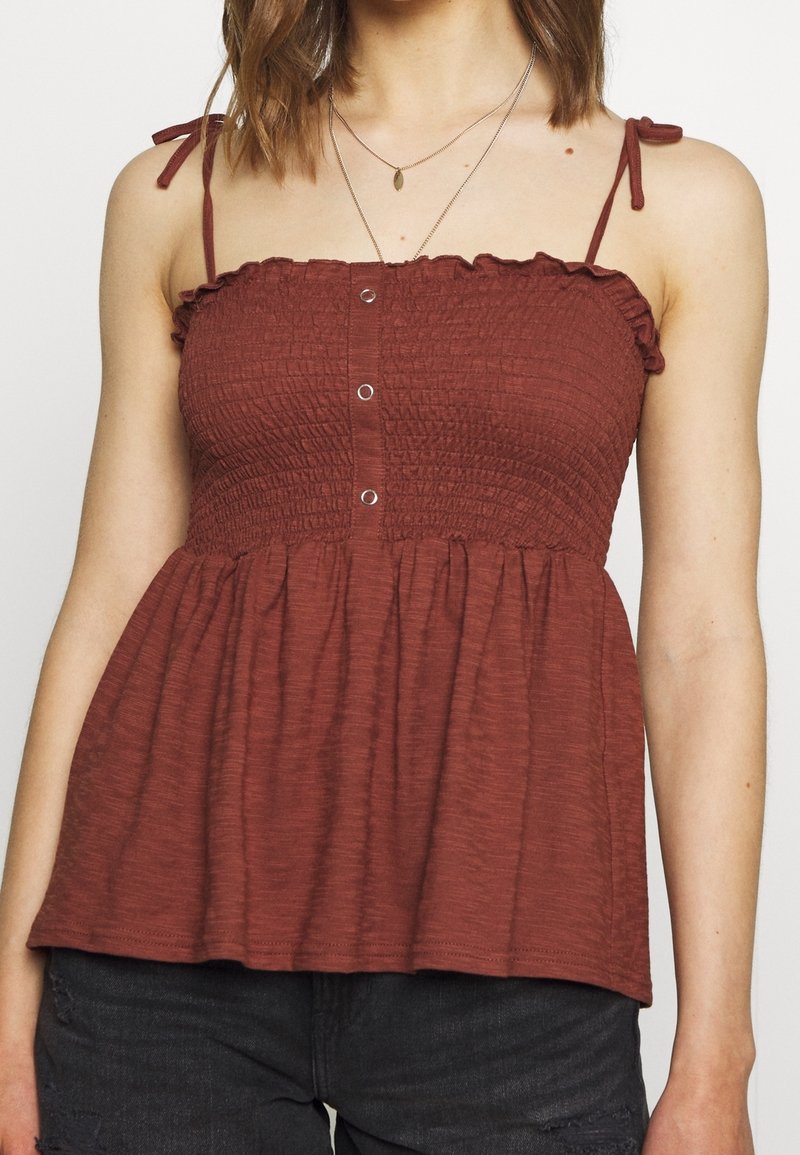 Haut sans manches texturé de couleur rouille rouge, avec un col à volants et des bretelles réglables ; présente des boutons à l'avant et un corsage ajusté avec un ourlet évasé.