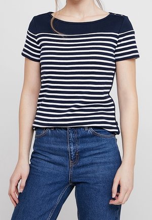 Femme portant un t-shirt à manches courtes rayé bleu marine et blanc, rentré dans un jean bleu taille haute, debout devant un fond uni.