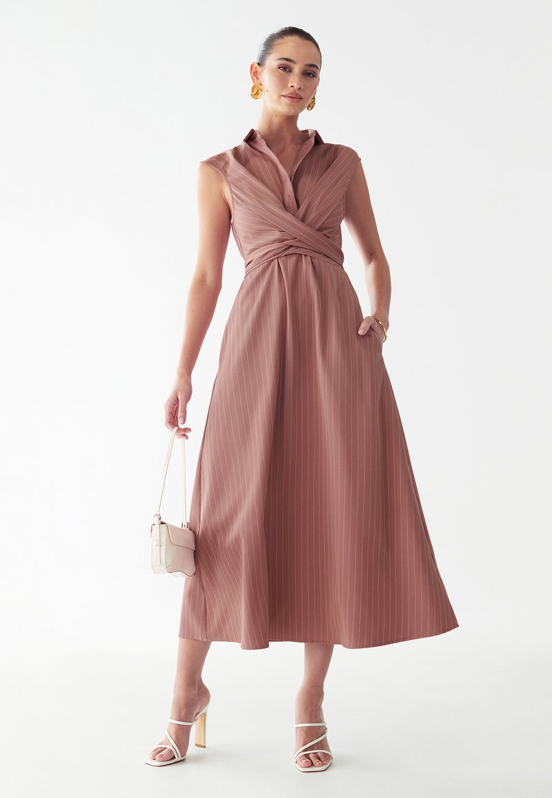 Mujer con vestido midi sin mangas de rayas finas en rosa polvo con cuerpo cruzado, sosteniendo un pequeño bolso blanco y sandalias blancas de tacón alto.