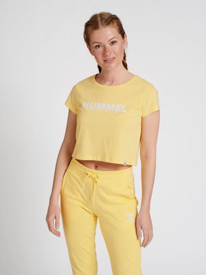 Ung kvinde med fletninger iført en gul crop top og matchende sweatpants med "Hummel" logo står mod hvid baggrund.